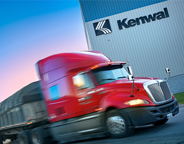 A red semi-truck hauling a large, covered load drives past the Kenwal Steel facility, with the Kenwal logo prominently displayed on the building’s exterior against a backdrop of a colorful sky at dusk.