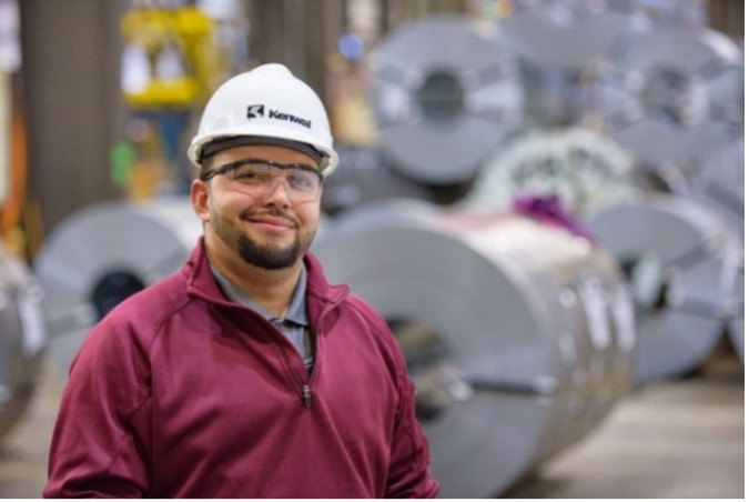 A smiling Kenwal staff member wearing their hard hat standing in front of several coils of flat rolled steel.