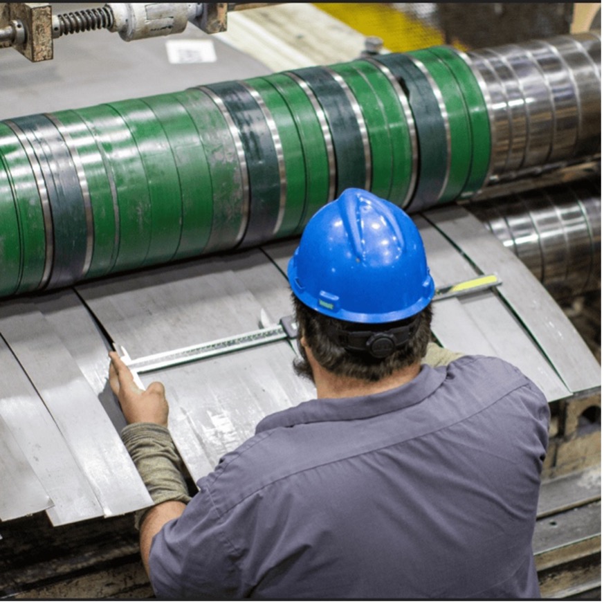 A Kenwal worker wearing a blue hard hat using handheld calipers to verify the precise width of steel strips exiting a heavy-duty slitting machine, demonstrating the manual quality control checks performed at a steel service center in Lebanon, TN.