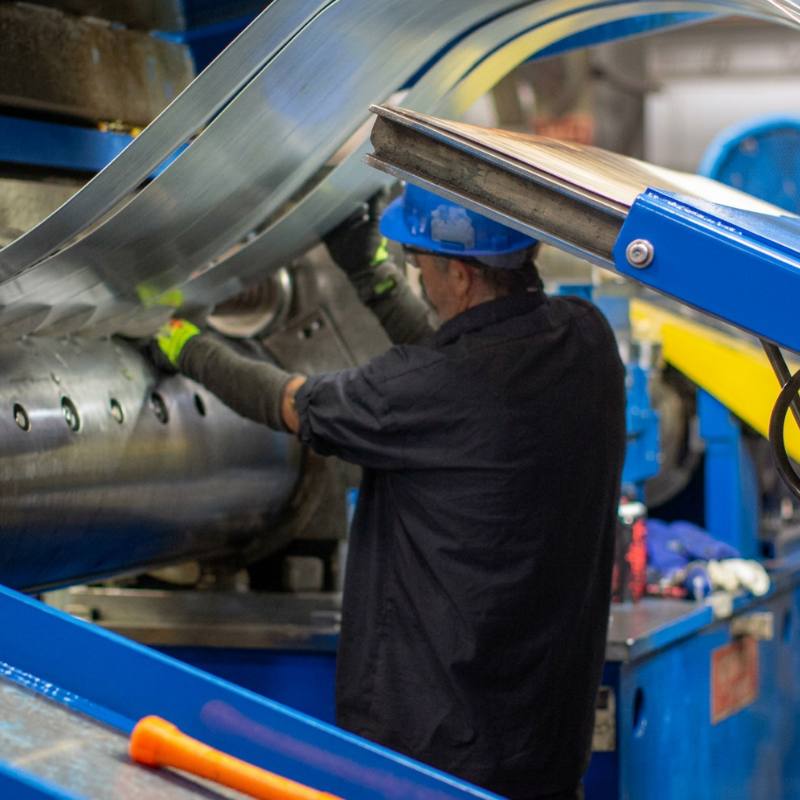 A worker in safety gear is operating a large metal‑processing machine with coils emerging as sliced strips. The composition highlights on‑site coil processing and operator interaction, relevant to reliable steel supply.