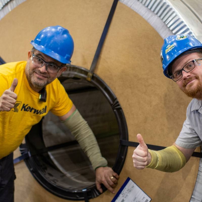 An industrial scene showing two workers in safety gear giving thumbs up beside a wrapped steel coil with visible markings; the composition emphasizes coil handling and workplace safety.