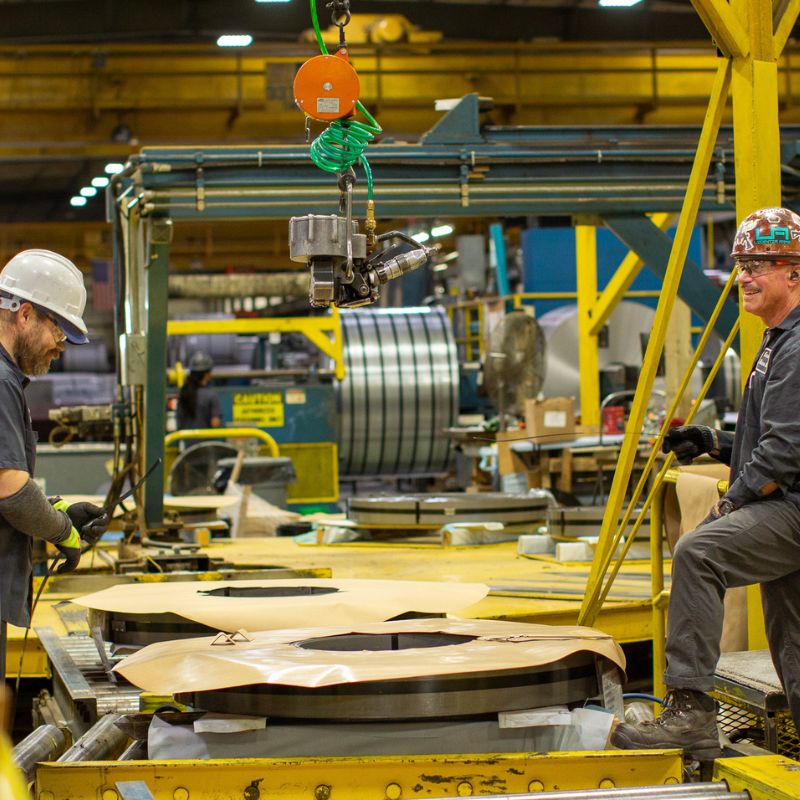 Kenwal-2026-09-steel-workers-packaging-coil Two Kenwal Steel workers smiling while packaging a flat-rolled steel coil in their manufacturing facility.